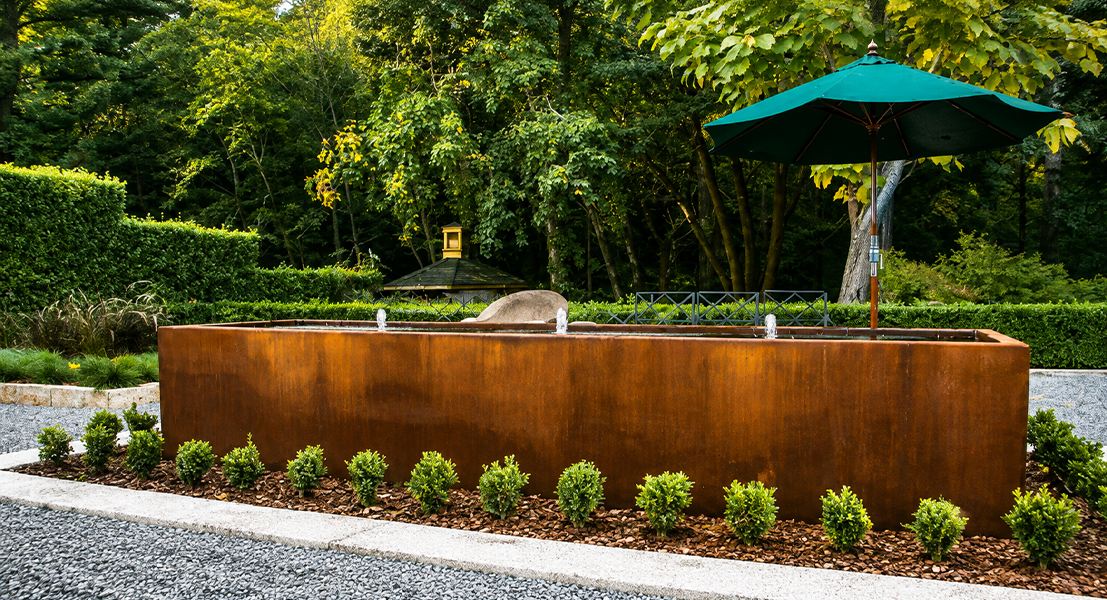 Grand bac rectangulaire monumental en acier Corten intégré dans un jardin paysager arboré.