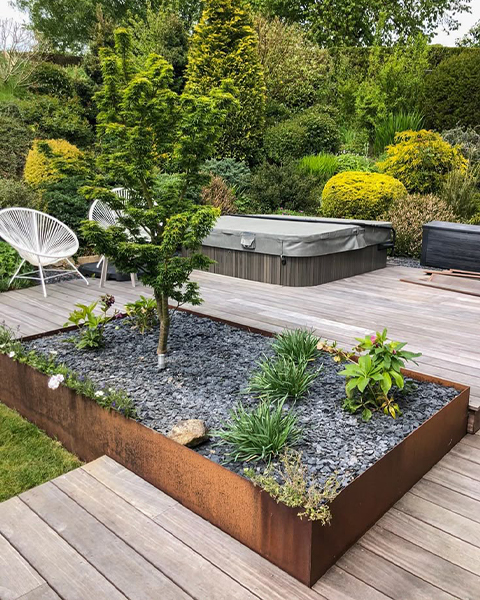 Grand bac paysager surélevé en acier Corten intégré à une terrasse en bois et planté d’un arbre et de végétaux variés.