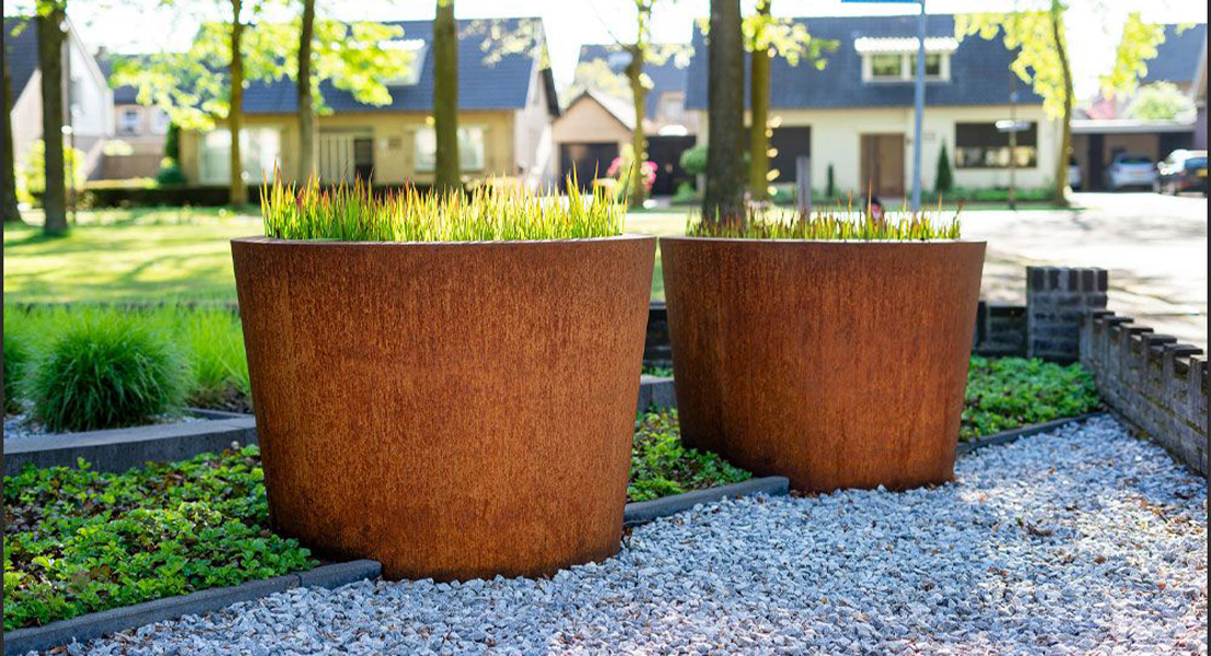 Deux jardinières rondes coniques en acier Corten plantées de graminées dans un jardin résidentiel.