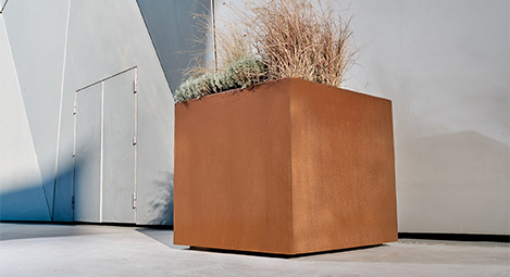 Jardinière Corten carrée et haute plantée de graminées devant une architecture contemporaine.