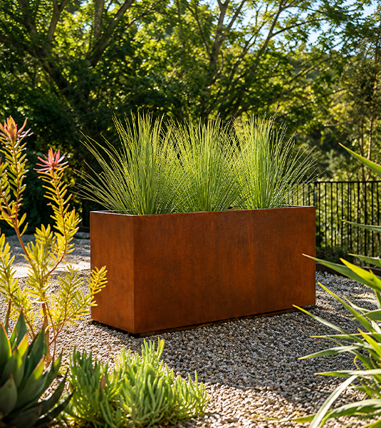 Jardinière Corten rectangulaire plantée de graminées dans un jardin paysager entouré de végétation.