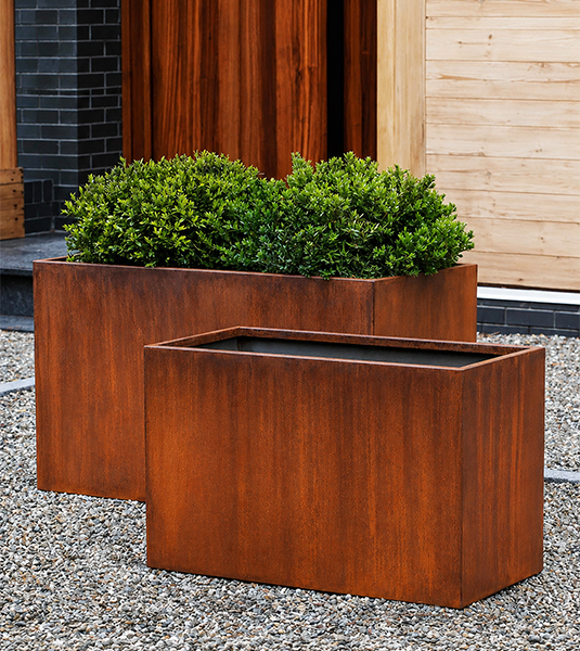 Deux jardinières Corten rectangulaires de tailles différentes accueillant des arbustes devant une maison contemporaine.